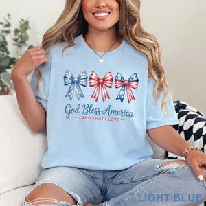 May include: Light blue t-shirt with the text "God Bless America - Land That I Love." The shirt features three bow designs in red, white, and blue, with stars and stripes patterns. The shirt is light blue.