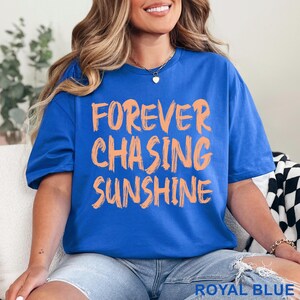 May include: A royal blue short-sleeved t-shirt featuring the text "FOREVER CHASING SUNSHINE" printed in a light orange, brush-stroke style font across the chest. The shirt has a crew neckline and a relaxed fit, with the colour labelled as royal blue.