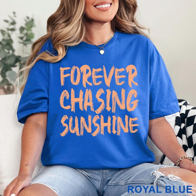 May include: A royal blue short-sleeved t-shirt featuring the text "FOREVER CHASING SUNSHINE" printed in a light orange, brush-stroke style font across the chest. The shirt has a crew neckline and a relaxed fit, with the colour labelled as royal blue.
