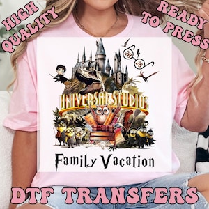 May include: A pink t-shirt featuring a colorful graphic design with the text "UNIVERSAL STUDIOS" and "Family Vacation". The design includes cartoon characters, a castle, and dinosaurs. The words "HIGH QUALITY" and "READY TO PRESS" are also visible.