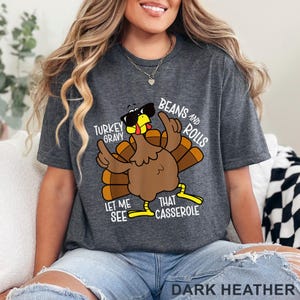 Puede incluir: Camiseta gris oscuro con un pavo de dibujos animados con gafas de sol y se&ntilde;alando hacia arriba. El texto en la camiseta dice "Turkey Gravy, Beans and Rolls, Let Me See That Casserole". Las palabras "Dark Heather" est&aacute;n impresas en la parte inferior.
