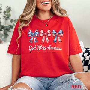 May include: Red t-shirt featuring three patriotic bow designs in red, white, and blue. The text "God Bless America" is printed below the bows. The shirt is a casual style, perfect for celebrating the USA.