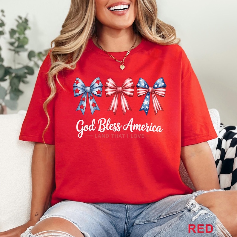 May include: Red t-shirt featuring three patriotic bow designs in red, white, and blue. The text "God Bless America" is printed below the bows. The shirt is a casual style, perfect for celebrating the USA.