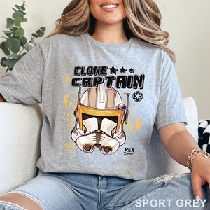 May include: Sport grey t-shirt featuring a detailed illustration of a clone trooper helmet with the text "CLONE CAPTAIN" above it. The design includes stars and a symbol, with the name "REX" below the helmet.