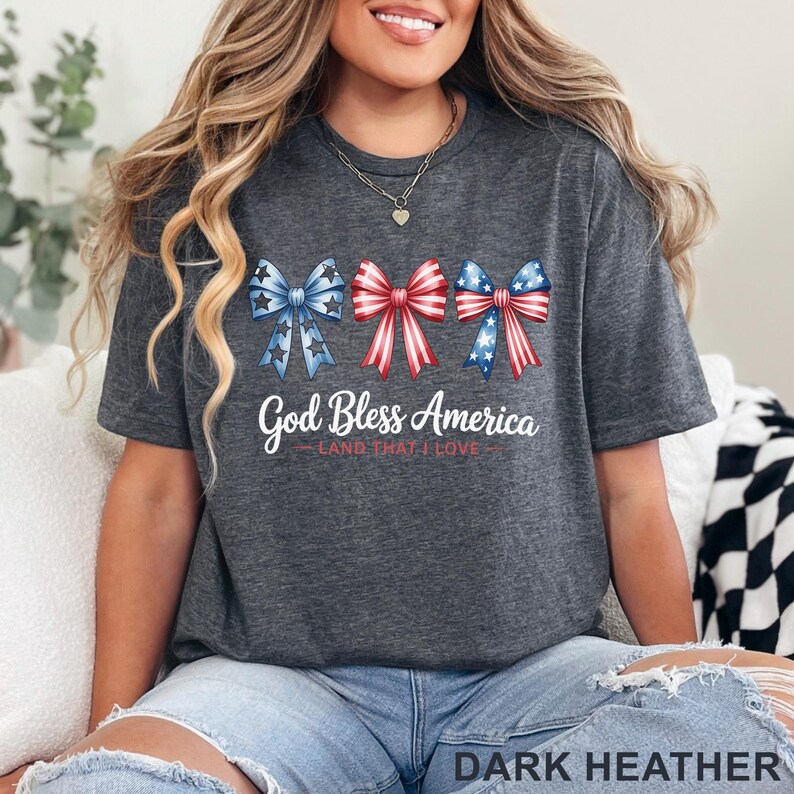 May include: Dark heather gray t-shirt featuring three patriotic bows in red, white, and blue. The shirt has the text "God Bless America - Land That I Love" in white script. The bottom of the shirt reads "DARK HEATHER".