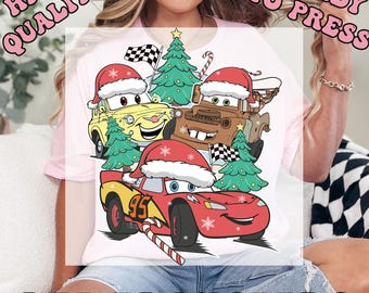 May include: A white t-shirt featuring a colorful graphic of cartoon cars wearing Santa hats and decorated with Christmas trees and candy canes. The text "HIGH QUALITY READY TO PRESS" and "DTF TRANSFERS" are printed on the image.