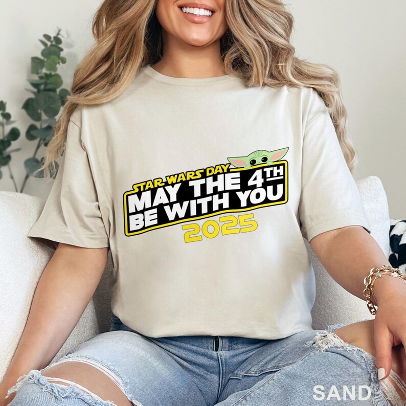 Puede incluir: Una camiseta color arena con el texto "STAR WARS DAY MAY THE 4TH BE WITH YOU 2025" en un dise&ntilde;o amarillo y blanco. El gr&aacute;fico incluye un personaje verde de Baby Yoda. La camiseta la lleva una persona.
