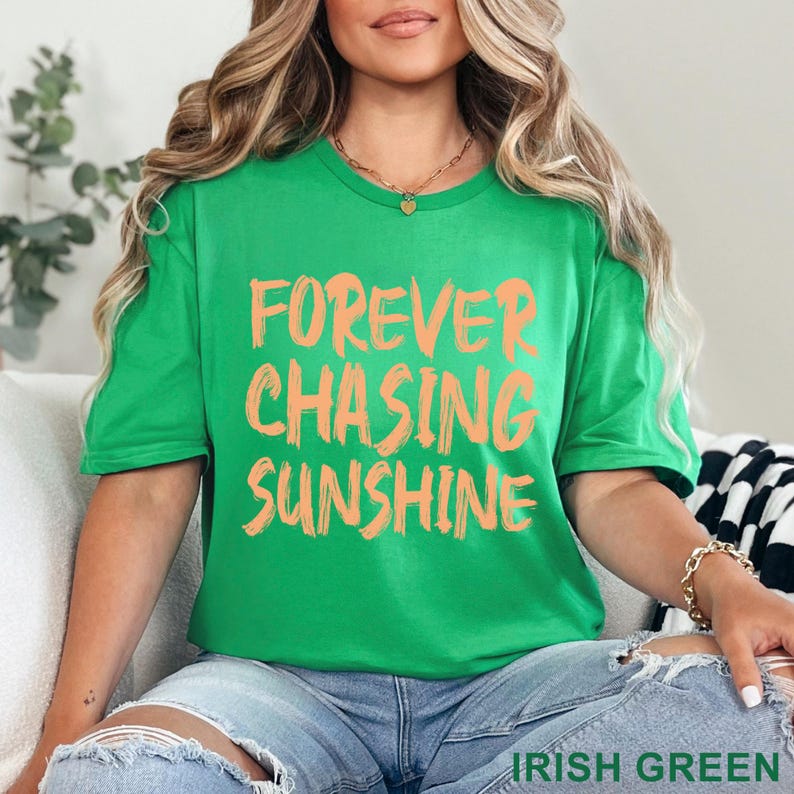 May include: A bright green short-sleeved t-shirt featuring the text FOREVER CHASING SUNSHINE printed in a bold, brush-stroke style font in a light orange colour. The shirt has a crew neckline and is worn with light blue denim jeans.