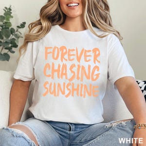 May include: A white short-sleeved t-shirt featuring the text FOREVER CHASING SUNSHINE printed in a bold, brushstroke-style peach-coloured font across the chest. The shirt has a crew neckline and a standard fit, paired with blue denim jeans.