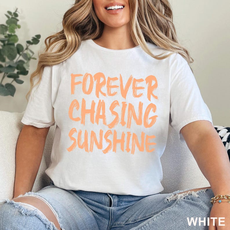 May include: A white short-sleeved t-shirt featuring the text FOREVER CHASING SUNSHINE printed in a bold, brushstroke-style peach-coloured font across the chest. The shirt has a crew neckline and a standard fit, paired with blue denim jeans.