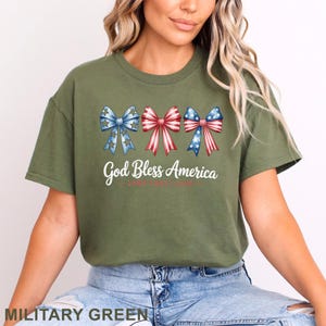 May include: Military green t-shirt with the text "God Bless America" and three patriotic bow designs in red, white, and blue. The bows have star and stripe patterns. The shirt is a comfortable, casual style.