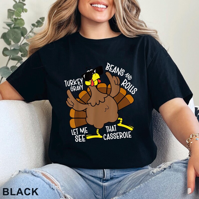 Puede incluir: Camiseta negra con un pavo de dibujos animados con gafas de sol y se&ntilde;alando hacia arriba. El texto en la camiseta dice: "Turkey Gravy, Beans and Rolls, Let Me See That Casserole."