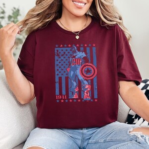 May include: Burgundy t-shirt featuring a distressed graphic of a superhero with a shield, set against a blue and red American flag design. The year "1941" is printed below the flag. The shirt has short sleeves and a crew neck.