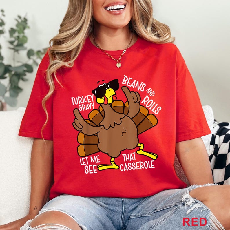 Puede incluir: Camiseta roja con un pavo de dibujos animados con gafas de sol y se&ntilde;alando hacia arriba. La camiseta tiene las frases "Turkey Gravy", "Beans and Rolls", "Let Me See" y "That Casserole" impresas. La palabra "RED" est&aacute; impresa en la parte inferior.