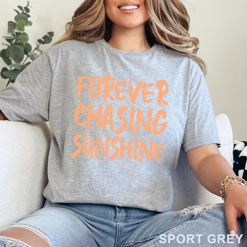 May include: A sport grey short-sleeved t-shirt featuring the text FOREVER CHASING SUNSHINE printed in a bold, light orange, brush-stroke style font across the chest. The shirt has a crew neckline and a standard fit, made of a heathered fabric.