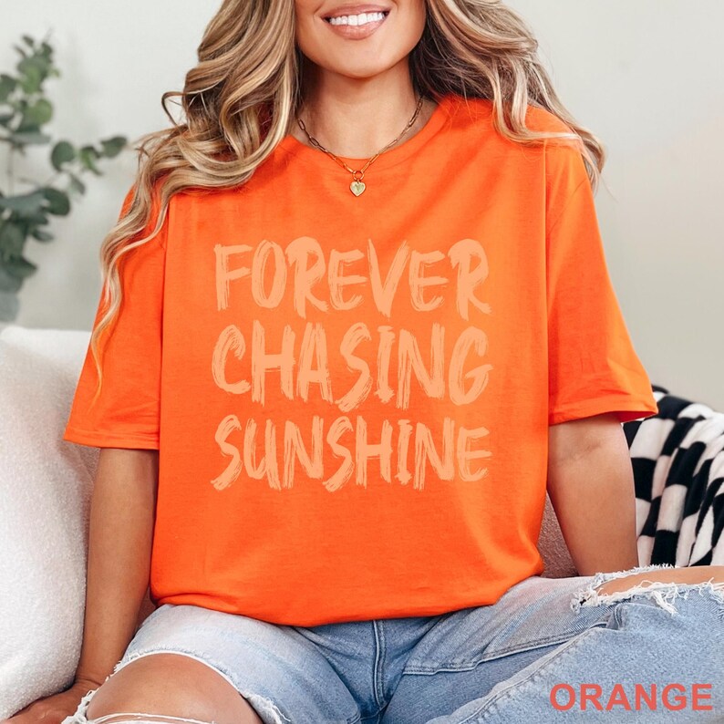 May include: A bright orange short-sleeved t-shirt featuring the text FOREVER CHASING SUNSHINE printed in a light peach, brush-stroke style font across the chest. The shirt has a crew neckline and is paired with light blue denim jeans.