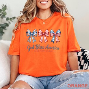 May include: Orange t-shirt with a patriotic design. The shirt features three bow illustrations in red, white, and blue, with stars and stripes. The text "God Bless America - Land That I Love" is printed below the bows.