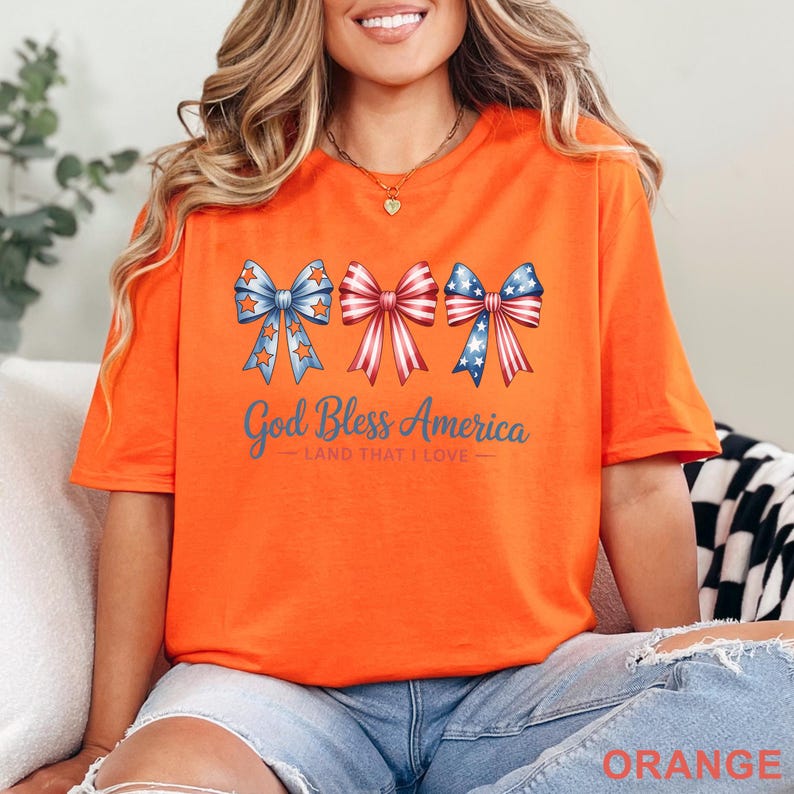 May include: Orange t-shirt with a patriotic design. The shirt features three bow illustrations in red, white, and blue, with stars and stripes. The text "God Bless America - Land That I Love" is printed below the bows.