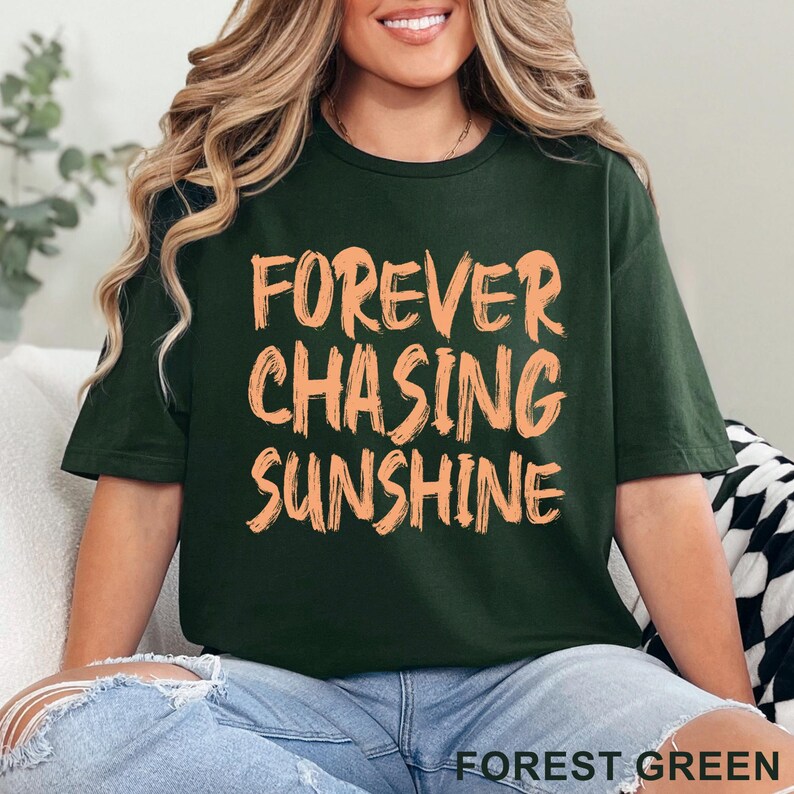 May include: A dark forest green short-sleeved t-shirt featuring the text "FOREVER CHASING SUNSHINE" printed in a light peach-coloured, brushstroke-style font across the chest. The shirt has a crew neckline and a standard, relaxed fit design.