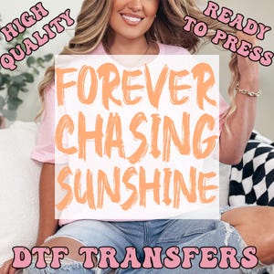 May include: A light pink t-shirt featuring the orange text FOREVER CHASING SUNSHINE. The image includes the text HIGH QUALITY and READY TO PRESS in pink, with DTF TRANSFERS at the bottom. The shirt has a crew neckline and short sleeves.