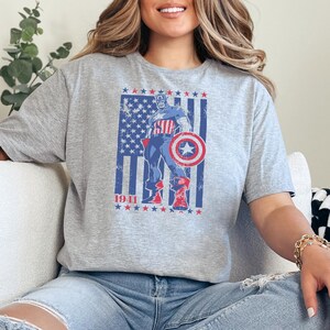 May include: A gray t-shirt featuring a graphic of Captain America with his shield, standing in front of an American flag design. The year "1941" is printed below the flag. The design is in shades of blue, red, and white.