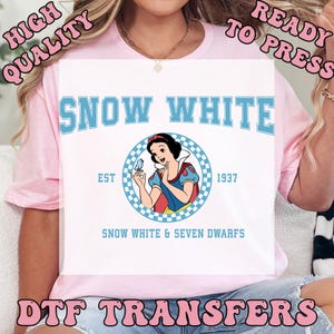 May include: A light pink t-shirt with a white graphic featuring the text "SNOW WHITE" in blue, with an illustration of Snow White holding a bird. The text "EST 1937" and "SNOW WHITE & SEVEN DWARFS" are also included. The words "HIGH QUALITY", "READY TO PRESS", and "DTF TRANSFERS" are also visible.