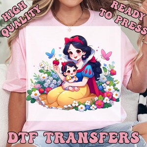 May include: A light pink t-shirt featuring a DTF transfer print of a cartoon Snow White holding a baby, surrounded by colorful flowers, butterflies, and a red apple. The text HIGH QUALITY READY TO PRESS DTF TRANSFERS is printed around the graphic.