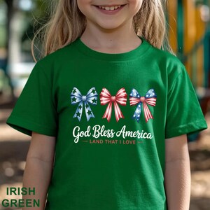 May include: Irish green t-shirt featuring three decorative bows in red, white, and blue, with the text "God Bless America - Land That I Love." The bows have star and stripe patterns.