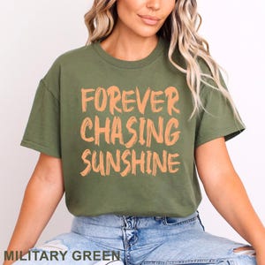 May include: An olive green short-sleeved t-shirt featuring the text FOREVER CHASING SUNSHINE printed in a light orange, brush-stroke style font across the chest. The shirt has a crew neckline and is paired with light blue denim jeans.