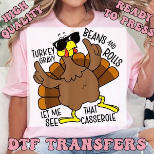Puede incluir: Una camiseta blanca con una ilustraci&oacute;n de un pavo de dibujos animados con gafas de sol y gesticulando con sus alas. El texto incluye "Turkey Gravy", "Beans and Rolls", "Let Me See" y "That Casserole". Tambi&eacute;n est&aacute;n las palabras "High Quality", "Ready to Press" y "DTF Transfers".