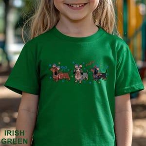 May include: Green t-shirt with a graphic of three dachshunds wearing patriotic headbands and bows. The dachshunds are brown, gray, and black. The shirt has the words "IRISH GREEN" printed at the bottom.