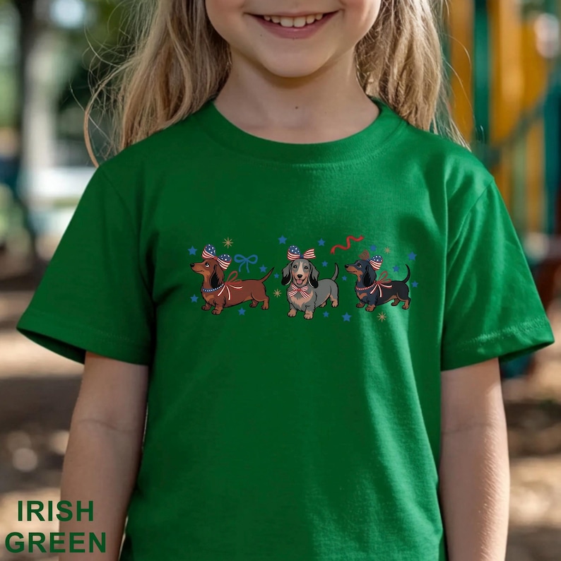 May include: Green t-shirt with a graphic of three dachshunds wearing patriotic headbands and bows. The dachshunds are brown, gray, and black. The shirt has the words "IRISH GREEN" printed at the bottom.