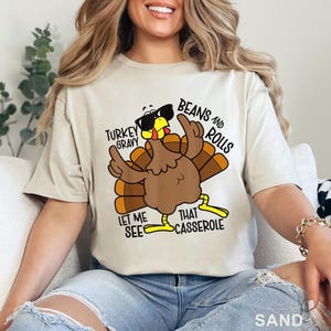 Puede incluir: Camiseta color arena con un pavo de dibujos animados con gafas de sol y se&ntilde;alando. El texto en la camiseta dice: "TURKEY GRAVY, BEANS AND ROLLS, LET ME SEE THAT CASSEROLE". El pavo es marr&oacute;n, amarillo y naranja.