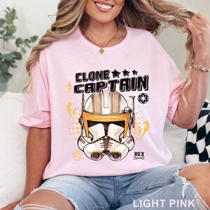 May include: Light pink t-shirt featuring a graphic of a Clone Captain helmet with the text "CLONE CAPTAIN" above it. The design includes stars, lightning bolts, and the name "REX". The shirt is made of a soft material.