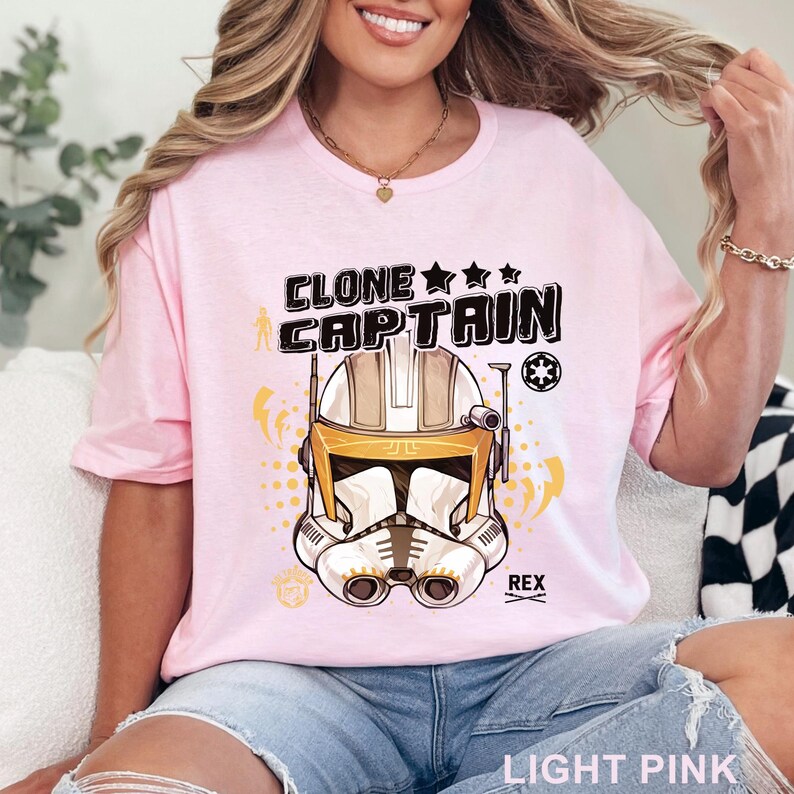 May include: Light pink t-shirt featuring a graphic of a Clone Captain helmet with the text "CLONE CAPTAIN" above it. The design includes stars, lightning bolts, and the name "REX". The shirt is made of a soft material.