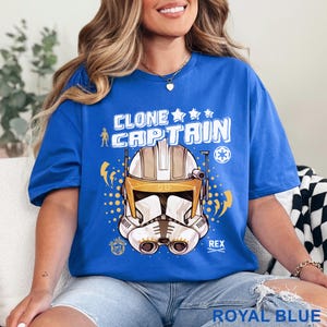 May include: Royal blue t-shirt featuring a graphic of a Clone Captain helmet with the text "CLONE CAPTAIN" above it. The design includes stars, lightning bolts, and the name "REX". The shirt is a solid royal blue color.