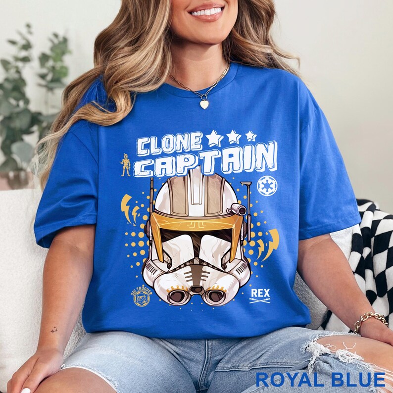 May include: Royal blue t-shirt featuring a graphic of a Clone Captain helmet with the text "CLONE CAPTAIN" above it. The design includes stars, lightning bolts, and the name "REX". The shirt is a solid royal blue color.