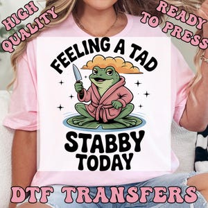 May include: A pink t-shirt with a graphic of a frog wearing a pink robe and holding a knife. The text on the shirt reads "FEELING A TAD STABBY TODAY". The words "HIGH QUALITY" and "READY TO PRESS" are also printed on the shirt.