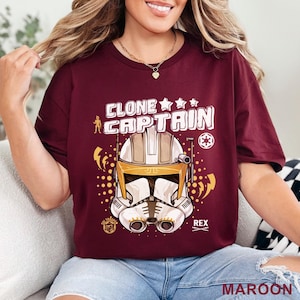 May include: Maroon t-shirt featuring a graphic of a Clone Captain helmet with the text "CLONE CAPTAIN" in white. The design includes stars, lightning bolts, and the name "REX". The word "MAROON" is printed at the bottom.