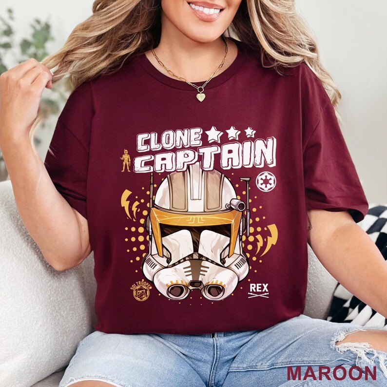 May include: Maroon t-shirt featuring a graphic of a Clone Captain helmet with the text "CLONE CAPTAIN" in white. The design includes stars, lightning bolts, and the name "REX". The word "MAROON" is printed at the bottom.