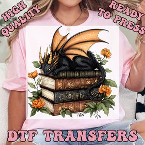 May include: A light pink t-shirt featuring a graphic print of a black dragon with orange wings resting on a stack of three antique books. The dragon is surrounded by orange flowers and green leaves. The text reads HIGH QUALITY READY TO PRESS DTF TRANSFERS.