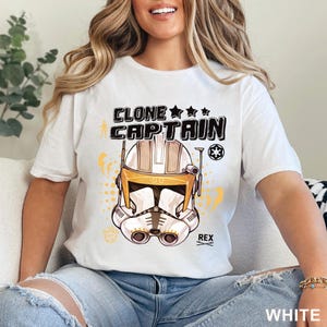May include: White t-shirt featuring a graphic of a Clone Captain helmet with the text "CLONE CAPTAIN" above it. The helmet is white, tan, and yellow. The word "REX" is below the helmet. The word "WHITE" is at the bottom right.