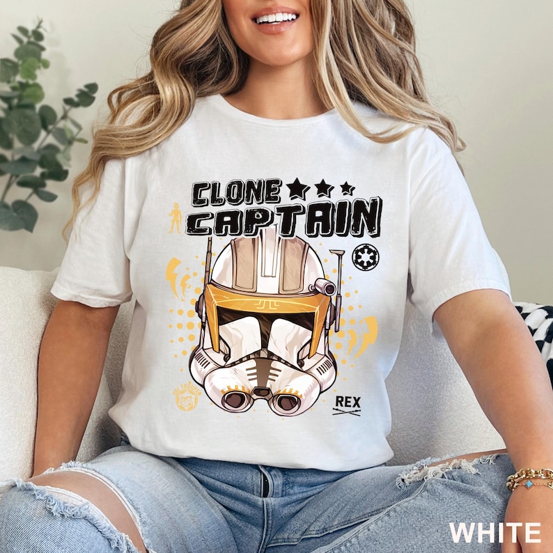 May include: White t-shirt featuring a graphic of a Clone Captain helmet with the text "CLONE CAPTAIN" above it. The helmet is white, tan, and yellow. The word "REX" is below the helmet. The word "WHITE" is at the bottom right.