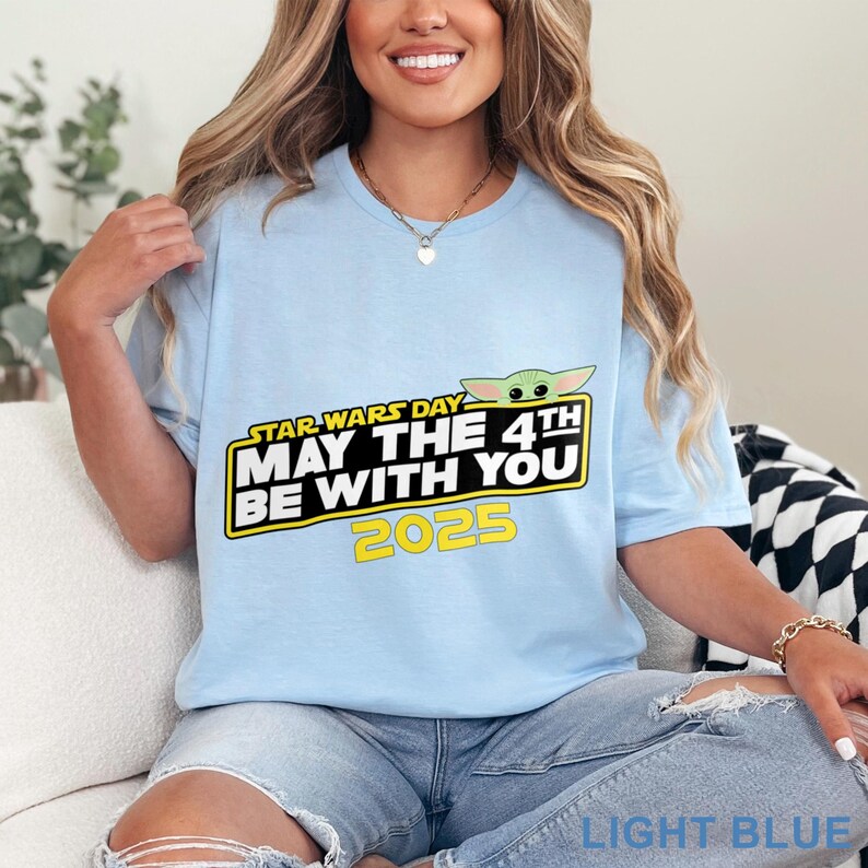 Puede incluir: Camiseta azul claro con el texto "STAR WARS DAY MAY THE 4TH BE WITH YOU 2025" en negro y amarillo. La camiseta tambi&eacute;n tiene un gr&aacute;fico de Grogu. La camiseta es de color azul claro.