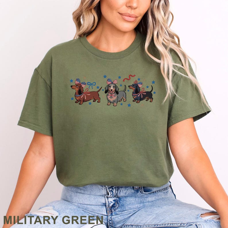 May include: Military green t-shirt featuring three dachshunds with patriotic bows and stars. The dogs are brown, gray, and black. The text "MILITARY GREEN" is at the bottom of the image.