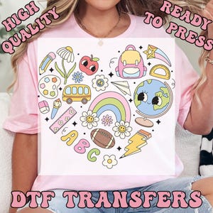 May include: A pink t-shirt featuring a heart-shaped design with school-themed illustrations. The design includes a school bus, rainbow, globe, and the letters ABC. The words "HIGH QUALITY READY TO PRESS DTF TRANSFERS" are also visible.