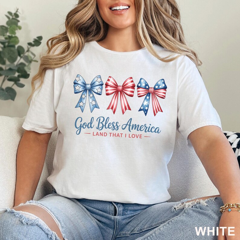 May include: White t-shirt featuring three decorative bows in red, white, and blue, with star accents. The text "God Bless America - Land That I Love" is printed below the bows. The word "WHITE" is printed at the bottom right.