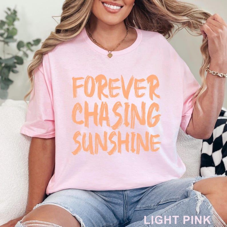 May include: A light pink t-shirt featuring the text FOREVER CHASING SUNSHINE printed in a bold, brush-stroke style orange font across the chest. The shirt has a crew neckline and short sleeves, with the words LIGHT PINK visible in the corner.