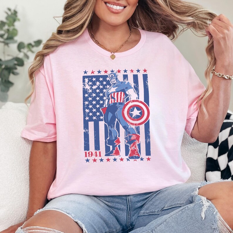 May include: A light pink t-shirt featuring a graphic of Captain America with a shield, standing in front of a distressed American flag design. The year "1941" is printed below. The shirt is a casual, round-neck style.