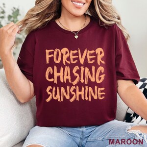 May include: A maroon short-sleeved t-shirt featuring the text "FOREVER CHASING SUNSHINE" printed in a light orange, brush-stroke style font across the chest. The shirt has a crew neckline and a standard fit, with the word MAROON visible at the bottom.